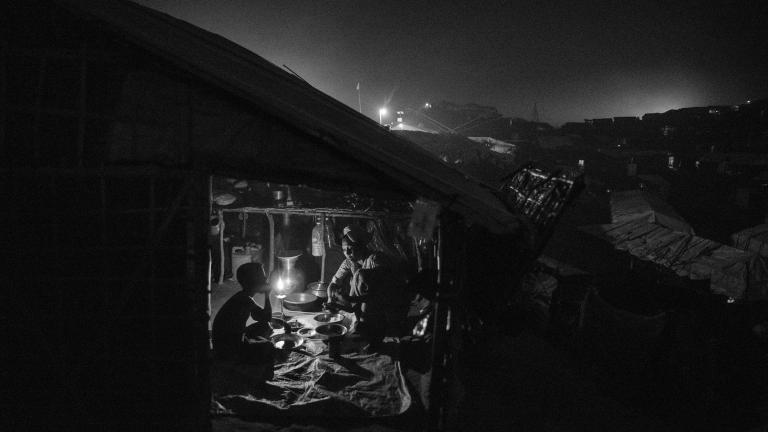 Une réfugiée rohingya sert de la nourriture à un garçon assis en face d’elle dans un abri sombre. L’intérieur est éclairé par une seule bougie.