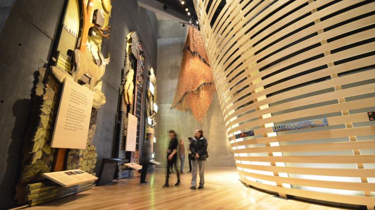 Quelques personnes dans une galerie de musée explorent de grands panneaux de bois décorés de figures d’animaux et d’œuvres perlées métisses. À droite, on voit des lattes de bois placées horizontalement et courbées. Visibilité masquée.