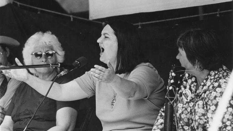 Photo en noir et blanc d’une femme chantant dans un micro, les bras levés. Une autre femme est assise à côté d’elle et la regarde.