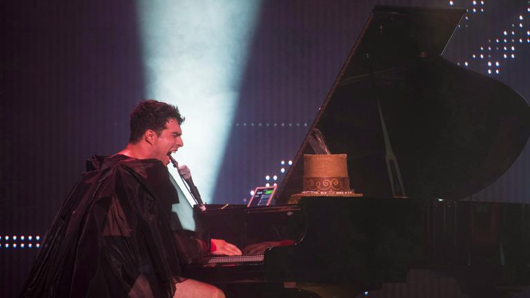 Un homme vêtu d’une cape noire diaphane et d’un short noir joue sur un piano à queue et chante intensément dans un micro. Derrière lui, on voit la lumière d’un projecteur dirigé vers le plafond.