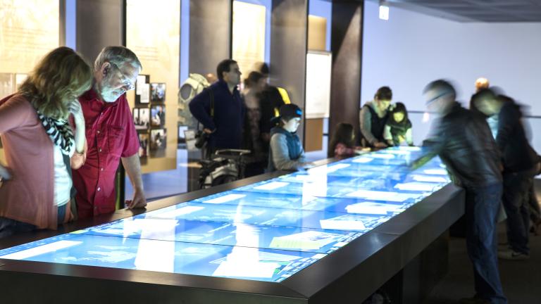 Des membres du public regardent une grande table rectangulaire dont la surface est un écran tactile.