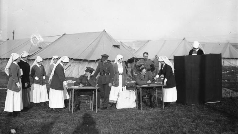 Photographie en noir et blanc d’infirmières et d’hommes en uniforme de soldats devant une série de tentes. À droite, une infirmière se tient derrière un écran de vote. Seules sa tête et ses épaules sont visibles.