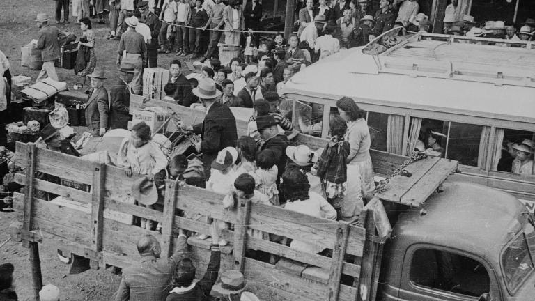 Une photographie en noir et blanc de personnes qui se font embarquer dans des camions alors que d’autres derrière elles font la queue.