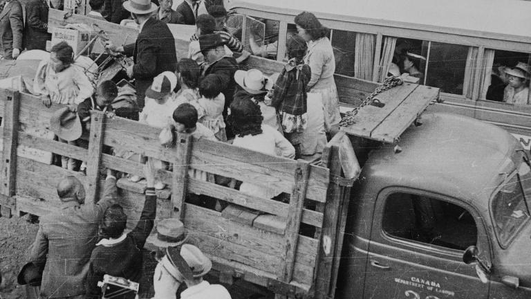 Une photographie en noir et blanc de personnes qui se font embarquer dans des camions alors que d’autres derrière elles font la queue.