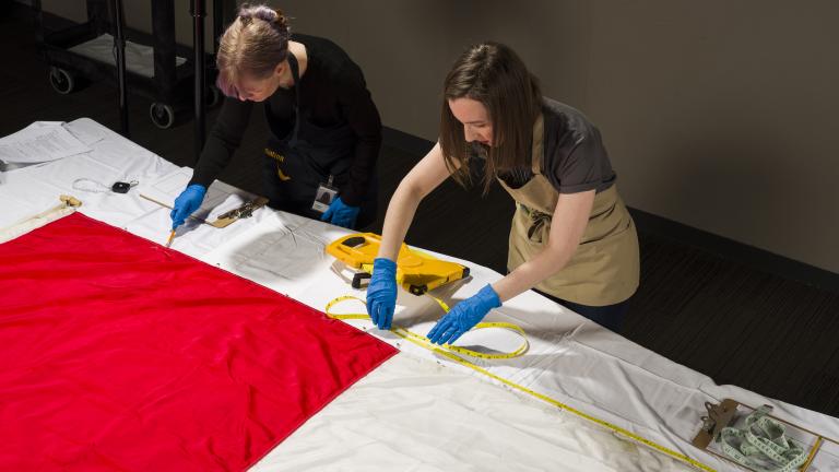 Deux femmes portant des gants et des tabliers de protection mesurent la longueur d’un grand drapeau du Canada, posé à plat sur une table.