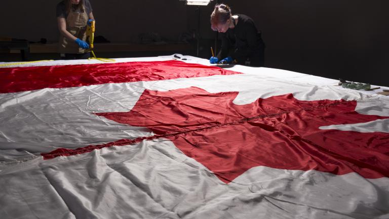 Deux femmes portant des gants et des tabliers de protection autour d’un grand drapeau du Canada, posé à plat sur une table.