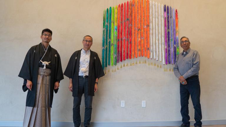 Trois hommes debout près d’une œuvre composée de mille grues multicolores en papier plié. Deux des hommes portent des vêtement traditionnels japonais. L’autre homme porte une chemise couleur denim et une cravate bolo.