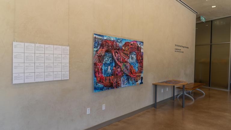 Une mosaïque de 24 tuiles sont suspendues à un mur près d’un panneau de texte. Les tuiles dépeignent des histoires sur les expériences de personnes métisses dans les systèmes scolaires coloniaux. Ensemble, les images forment un symbole de l’infini inspiré d’une ceinture fléchée rouge sur un fond bleu.