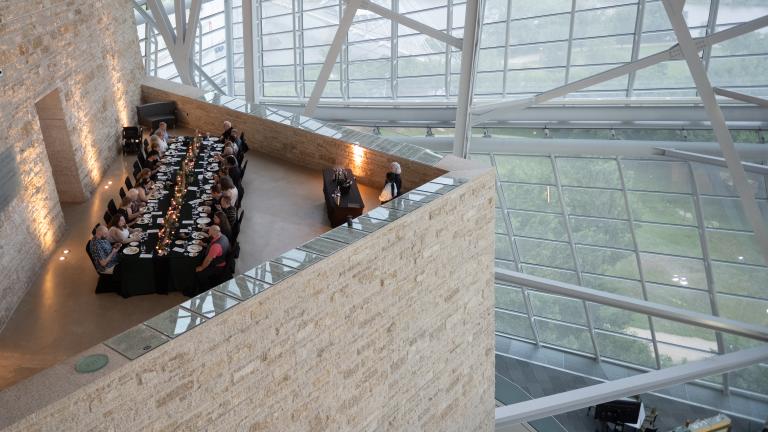 Une élégante table longue avec des personnes dînant dans un espace à l'architecture remarquable, doté de hautes fenêtres en verre et d'un plafond élevé soutenu par des poutres métalliques.