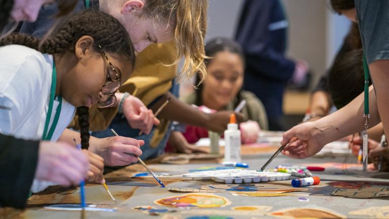 Un groupe d’enfants et de jeunes peint avec de petits pinceaux, de part et d’autre d'une longue table. Au premier plan, il y a des peintures de différentes couleurs. Visibilité masquée.