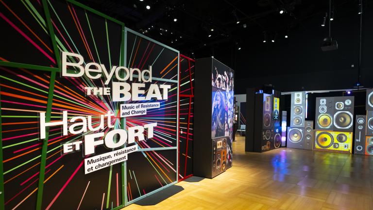 L’entrée d’une exposition muséale avec un panneau graphique comprenant des rayons colorés et le texte « Haut et fort : Musique, résistance et changement » en anglais et en français. Une image projetée d’une pile de divers haut-parleurs couvre le mur adjacent. Visibilité masquée.