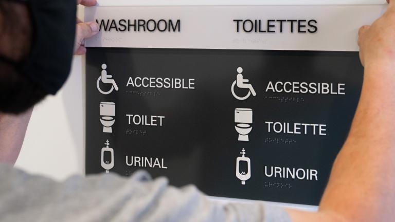 A man holds up a sign that says "Washroom" as it is mounted to a wall. Underneath the title, the words "accessible," "toilet" and "urinal" are accompanied by descriptive icons. Visibilité masquée.