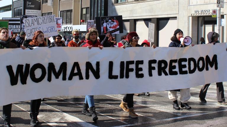 Six personnes portent une longue bannière blanche sur laquelle on peut lire les mots anglais en lettres majuscules noires "FEMME VIE LIBERTÉ" dans une rue de la ville de Toronto. Derrière elles, un défilé de femmes porte d'autres pancartes de protestation pour la Journée internationale des femmes. Visibilité masquée.