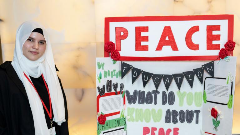 Dans une galerie de musée, une étudiante se tient à côté d’une table présentant le projet qu’elle a créé. Le projet porte le mot « paix » en grosses lettres. Visibilité masquée.