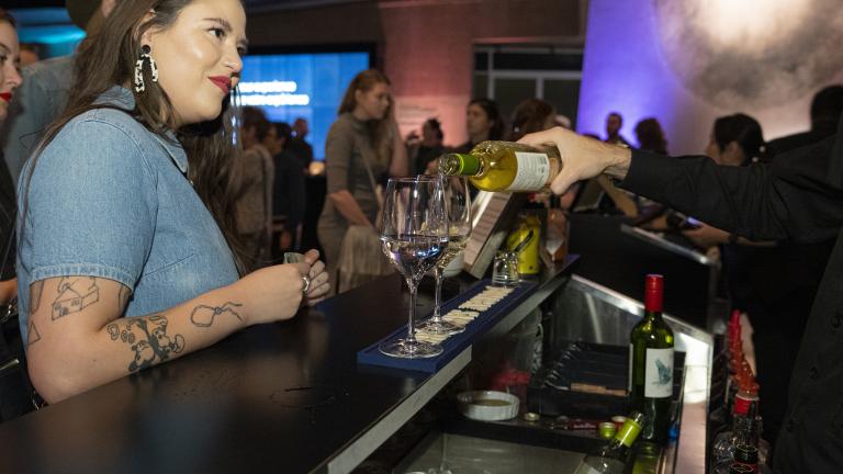A woman waits for a drink at a bar in a museum space, while a bartender pours glasses of wine. Visibilité masquée.