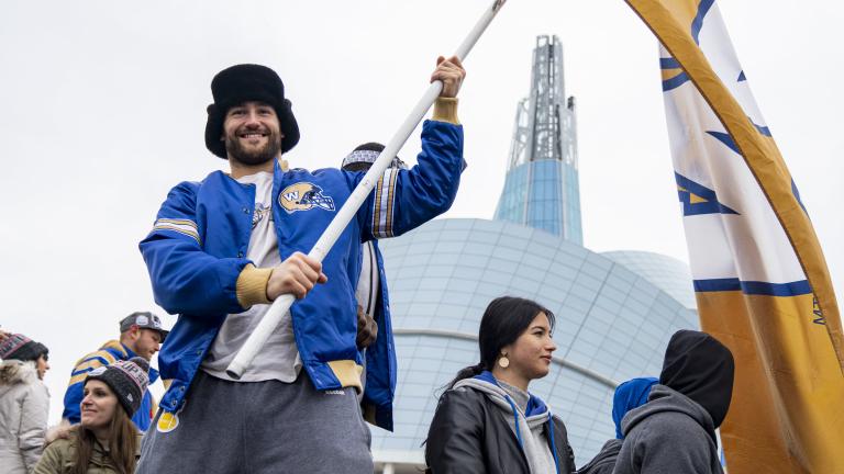 Un homme souriant dans une foule, tenant un grand drapeau jaune avec un « W » bleu et blanc au centre. L’homme porte une veste bleue des Blue Bombers de Winnipeg et se tient devant le Musée canadien pour les droits de la personne. Visibilité masquée.