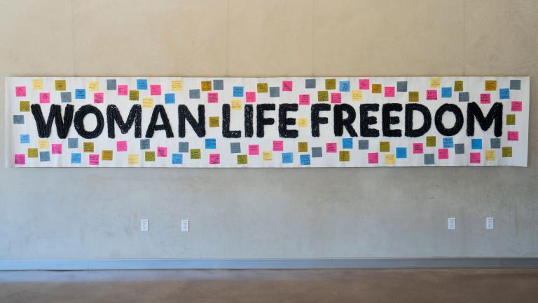Une banderole blanche est accrochée à un mur, portant les mots "Femme, Vie, Liberté" en noir. De petits carrés de couleur avec du texte sont attachés à la bannière autour du texte. Visibilité masquée.