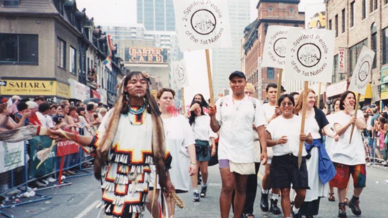 Un groupe de personnes portant des pancartes sur lesquelles on peut lire « 2-Spirited People of the 1st Nations » marchent au centre d’une rue de la ville, tandis que des gens regardent derrière des barrières de part et d’autre de la rue. Visibilité masquée.
