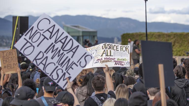 Une foule de personnes faisant dos à la caméra. Certains membres de la foule tiennent des pancartes au-dessus de leur tête qui disent, en anglais : « Je ne peux pas respirer #BLM », « Le Canada n'est pas à l'abri du racisme », et « Justice pour tous : mettez fin au racisme violent, subtil et systémique maintenant!!! ». Visibilité masquée.