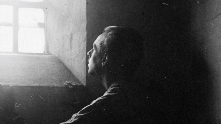 Un homme se tient dans l’ombre d’une cellule de pierre austère, regardant vers le haut la lumière vive du soleil qui entre par une fenêtre à barreaux. Visibilité masquée.