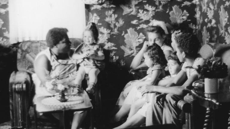 Une image en noir et blanc de femmes et de jeunes enfants assis sur des sofas et des chaises dans un petit salon. Visibilité masquée.