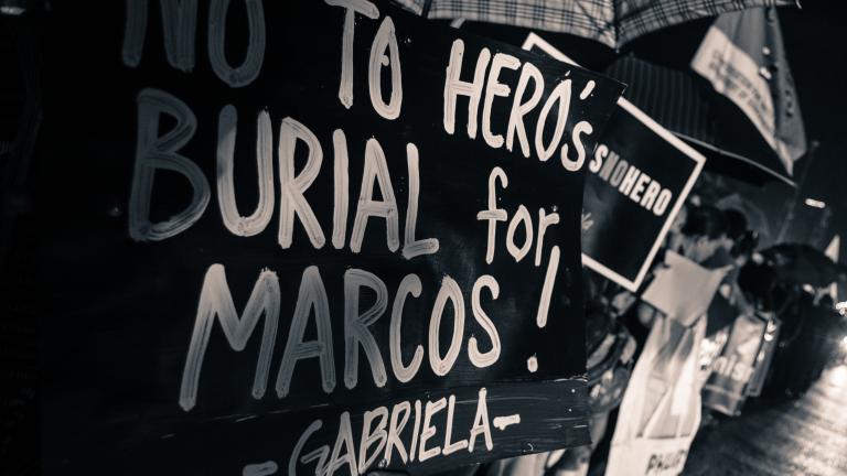 Des personnes debout sous des parapluies au bord d’une route. Les phares des véhicules qui les dépassent sont visibles sur le côté droit de l’image. Au premier plan à gauche, une pancarte leur cache le visage et indique « Non à un enterrement de héros pour Marcos! Gabriela. » Visibilité masquée.