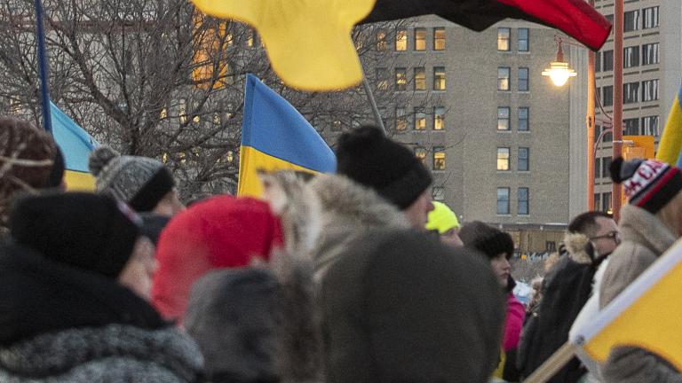 Une foule de personnes emmitouflées se tient debout dans une rue de ville pendant l’hiver, tenant de multiples drapeaux ukrainiens bleus et jaunes ainsi qu’un drapeau canadien. Le rassemblement semble être une manifestation en appui à l’Ukraine. De grands édifices à bureaux et des feux de circulation sont à l’arrière-plan sous un ciel couvert. Visibilité masquée.