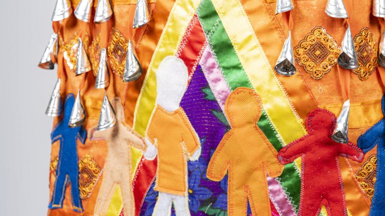 Une section d'une robe orange à clochettes est représentée. Les clochettes, ou petits pendentifs argentés en forme de cône, sont visibles en haut de l'image. Sous elles, sept figures humaines sont représentées, toutes de couleurs et de tailles différentes. L'une des figures porte une chemise orange. Ces figures humaines se tiennent la main à l'intérieur d'un tipi multicolore. Visibilité masquée.