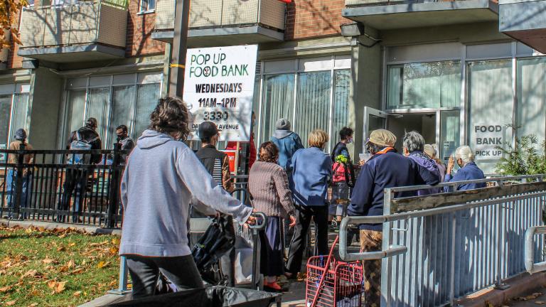 Une file de personnes attend à l’extérieur d’un bâtiment. Des panneaux indiquent qu’il s’agit d’une banque alimentaire. Un drapeau canadien flotte au-dessus d’elles. Visibilité masquée.