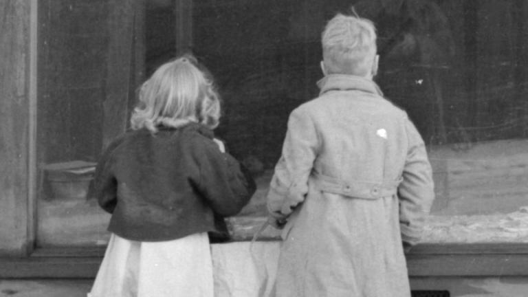Photographie en noir et blanc de deux enfants regardant la vitrine d’un magasin abandonné. Visibilité masquée.