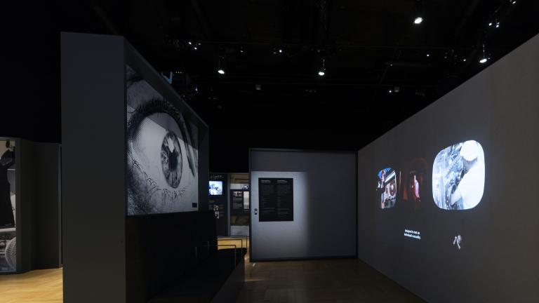 Un panneau d’exposition à gauche présente l’image d’un grand œil. À droite, trois petites vidéos sont projetées sur le mur. L’espace est sombre.