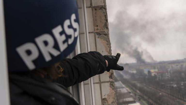 Une personne portant un casque bleu foncé marqué du mot « PRESS » montre, depuis la fenêtre d’un balcon surplombant une route, une colonne de fumée à la suite d’une explosion. Visibilité masquée.