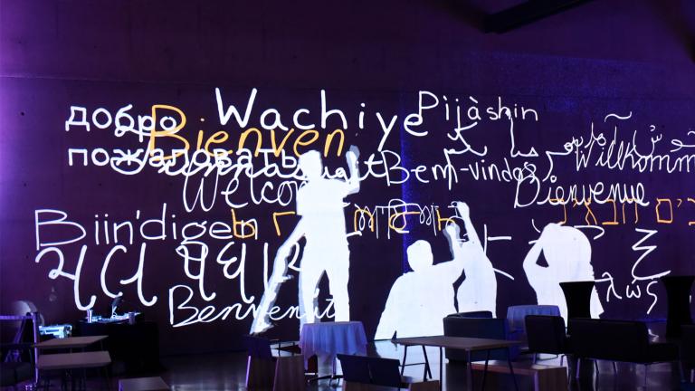 Image fixe d’une animation montrant des silhouettes humaines écrivant « bienvenue » en plusieurs langues en blanc et jaune sur un mur violet. Visibilité masquée.