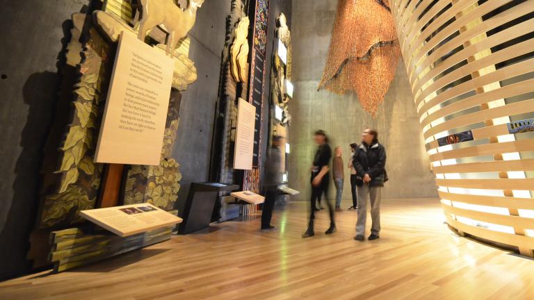 Cinq personnes regardent des expositions imposantes dans la galerie « Perspectives autochtones » du Musée. La plus proche est en bois et représente des arbres, des animaux et une plaque. Derrière les personnes, on aperçoit un théâtre de forme circulaire, construit avec des lattes de bois courbées. Visibilité masquée.