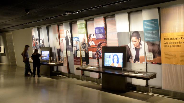 Deux personnes interagissent avec l’un de deux écrans vidéo dans une galerie de musée. Il y a des images de gens et des blocs de texte en couleur sur le mur.