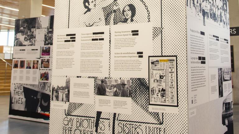 Exposition dans un espace moderne présentant des affiches historiques et des textes sur le militantisme 2ELGBTQ+, avec des personnes observant l’exposition et une personne sur un balcon au-dessus.