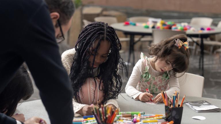 Deux enfants sont assis à une table et font de l’art tandis qu'un adulte s'appuie sur la table pour les observer. Visibilité masquée.