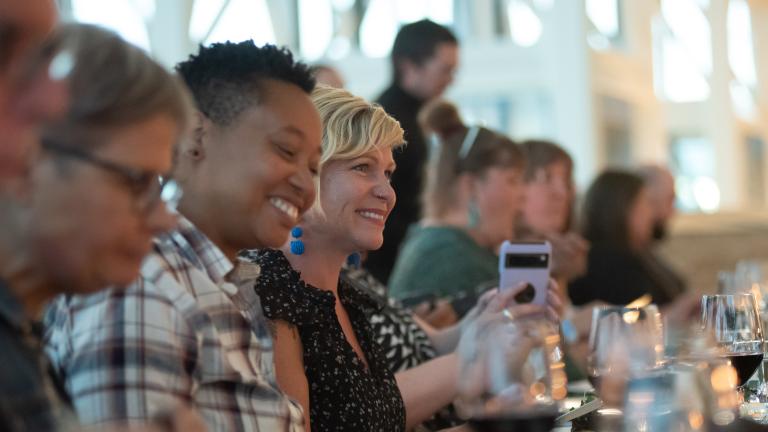 Un groupe de personnes est assis à une longue table, riant et appréciant un repas ensemble. L’accent est mis sur deux personnes qui sourient joyeusement – l’une portant une chemise à carreaux et l’autre un haut noir à motifs, avec des verres et des assiettes soigneusement disposés devant elles. Visibilité masquée.