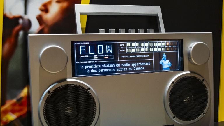 Grande radio de type "boombox" avec un écran en haut qui affiche "FLOW", des sous-titres et un interprète ASL. Visibilité masquée.