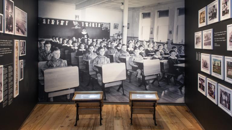 Une exposition muséale qui comprend de petits pupitres d’école devant un grand fond photographique d’enfants autochtones assis à leur pupitre dans une classe. Visibilité masquée.