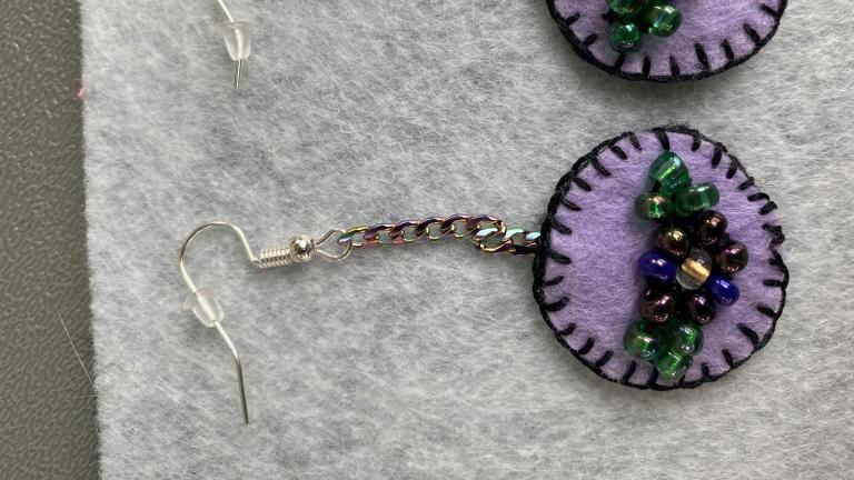 Une paire de boucles d'oreilles en feutre violet clair avec des bordures cousues violet foncé. Au milieu des boucles d'oreilles se trouvent des fleurs en perles. Visibilité masquée.