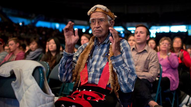 Un homme âgé vêtu d'une chemise à carreaux et d'accessoires autochtones traditionnels est assis dans une foule. Il a les bras pliés aux coudes et les mains levées, ouvertes l’une face à l’autre.