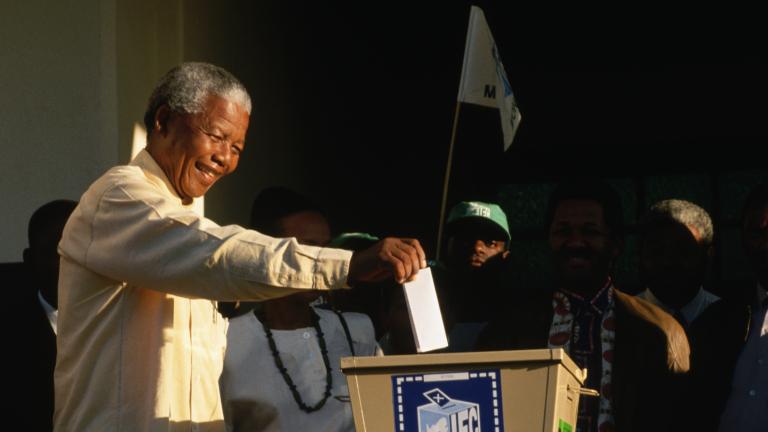  Nelson Mandela étendant son bras droit pour déposer un papier plié dans une boîte de scrutin. Quelques personnes sont debout autour de lui.