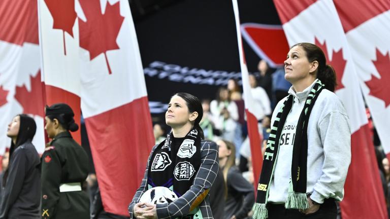 Des personnes se tiennent debout sur le terrain d’un stade pendant une cérémonie d’avant-match, tandis que de grands drapeaux canadiens sont brandis derrière elles, remplissant l’arrière-plan de rouge et de blanc. Visibilité masquée.