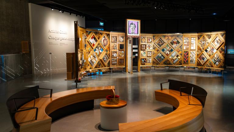 Exposition muséale présentant un grand mur de bois incurvé et autoportant sur lequel sont fixés de nombreux petits objets disposés en losanges et en rectangles. Au premier plan, des bancs circulaires créent un espace accueillant où les gens peuvent regarder et contempler l’exposition.