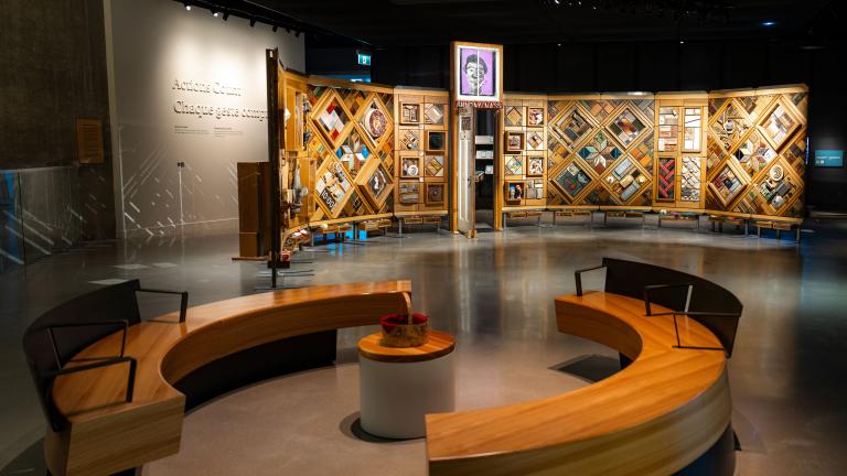 Exposition muséale présentant un grand mur de bois incurvé et autoportant sur lequel sont fixés de nombreux petits objets disposés en losanges et en rectangles. Au premier plan, des bancs circulaires créent un espace accueillant où les gens peuvent regarder et contempler l’exposition. Visibilité masquée.
