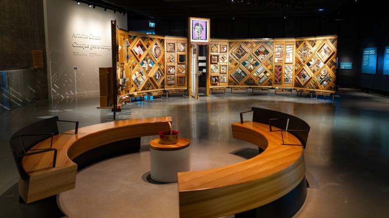 Exposition muséale présentant un grand mur de bois incurvé et autoportant sur lequel sont fixés de nombreux petits objets disposés en losanges et en rectangles. Au premier plan, des bancs circulaires créent un espace accueillant où les gens peuvent regarder et contempler l’exposition. Visibilité masquée.