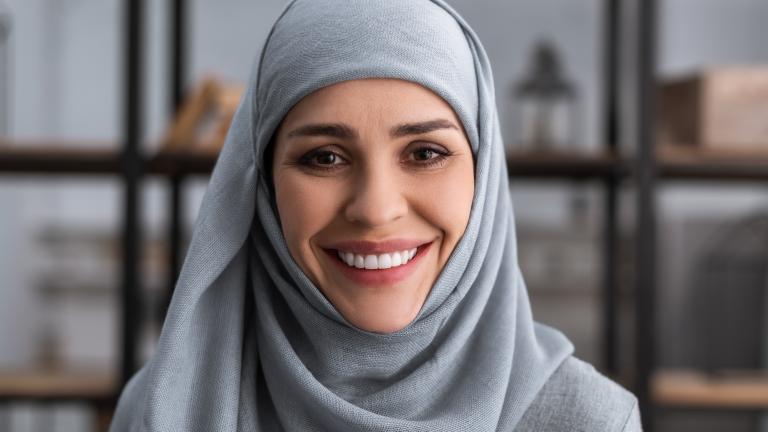 Une femme portant un hidjab bleu sourit vers l'objectif. Il y a une étagère de livres en arrière-plan. Visibilité masquée.