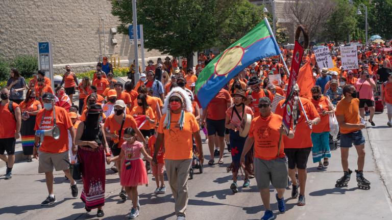 Des centaines de personnes vêtues de chemises orange, dont beaucoup portent également des articles traditionnels autochtones comme une coiffe ou une jupe à rubans et brandissent des pancartes, défilent dans une large rue pavée. Visibilité masquée.