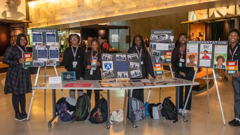 Dans une salle de musée, six étudiants noirs présentent fièrement leur travail sur des affiches et sur une table. Visibilité masquée.